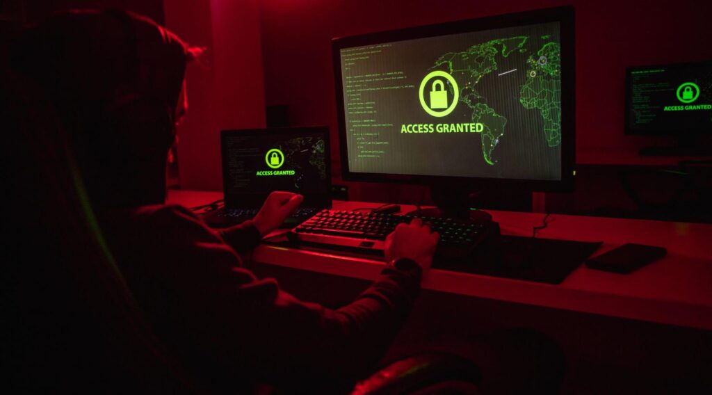 An anonymous hacker is stealing data on a computer in red lighting.