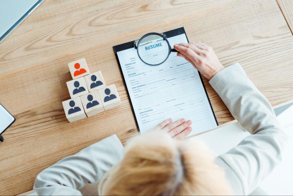 Top view of a blonde recruiter reviewing a resume with a magnifying glass beside wooden blocks with human icons.
