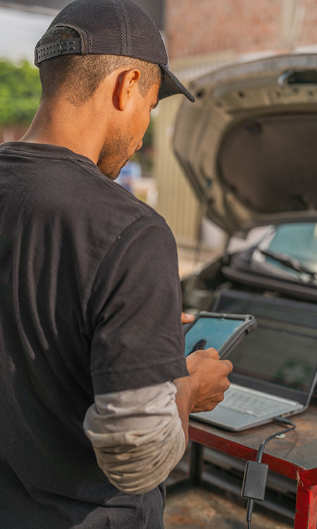 A person stands by a car with its hood open, using a tablet while a laptop rests on a nearby table.