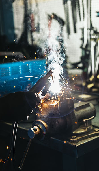 A hand holding welding pliers sparks and smoke erupting from a metal object in a workshop, surrounded by tools and equipment.