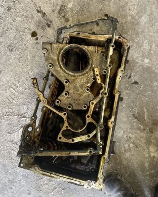 An oil-covered engine block with a disassembled oil pan and gasket, resting on a concrete floor.