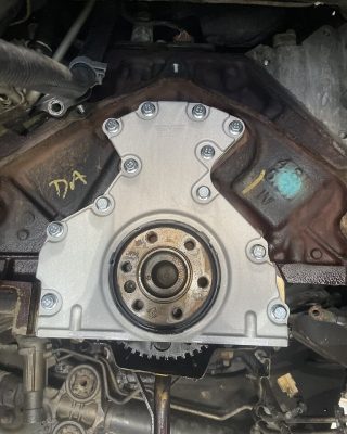 Close-up of an engine's timing cover, featuring a metallic design with bolts and a gear, set against a dark engine block background.