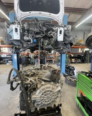A car engine is being lifted by a hoist in a garage, with tools and equipment visible in the background.