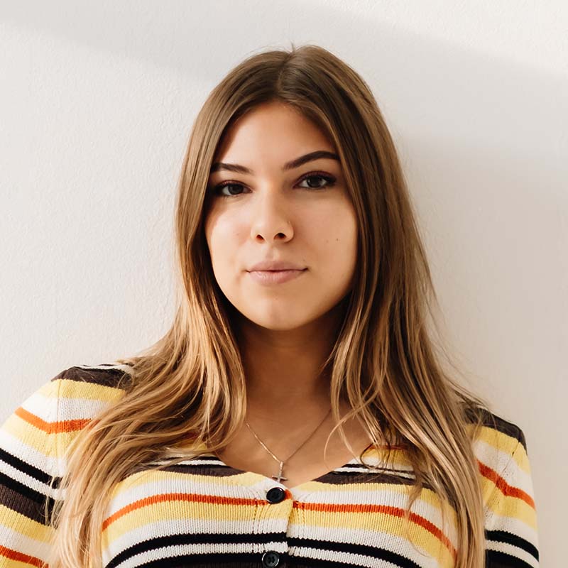 Woman looking at the camera with long brown hair in a black, white, yellow and orange striped sweater