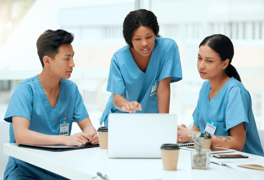 Three nurses in blue scrubs gathered around a laptop, discussing the study materials in a bright break room.