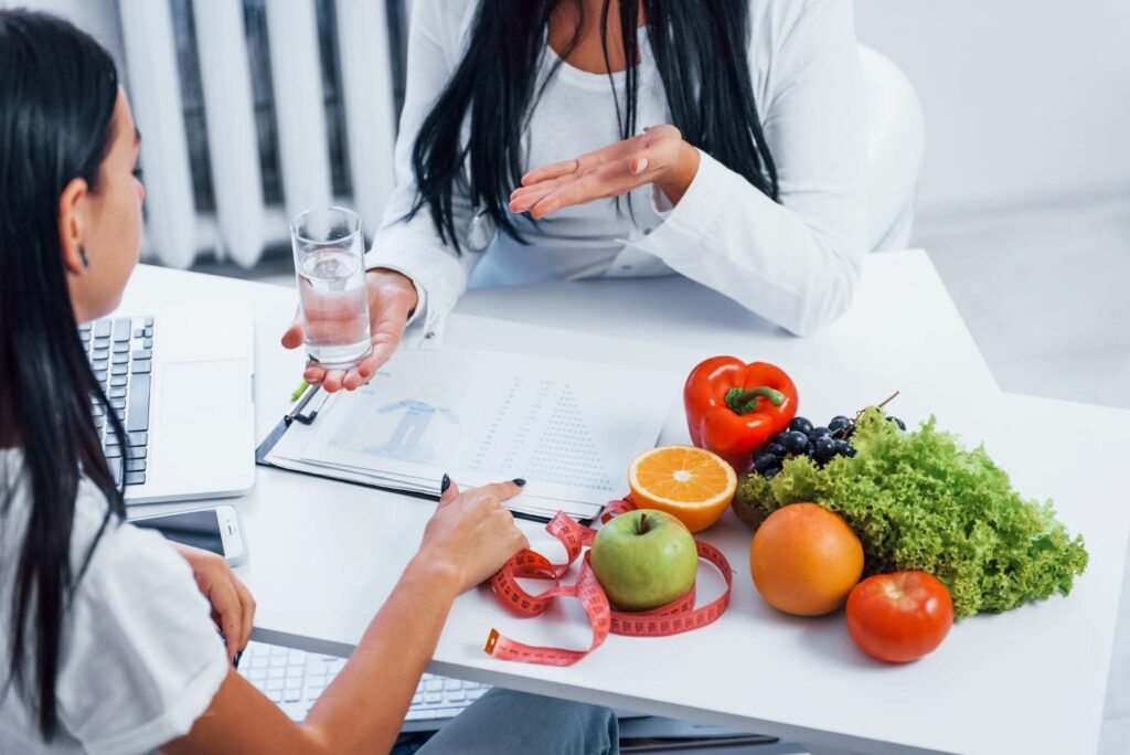 Woman holding a glass of water next to an assortment of fruits and vegetables across from a woman.