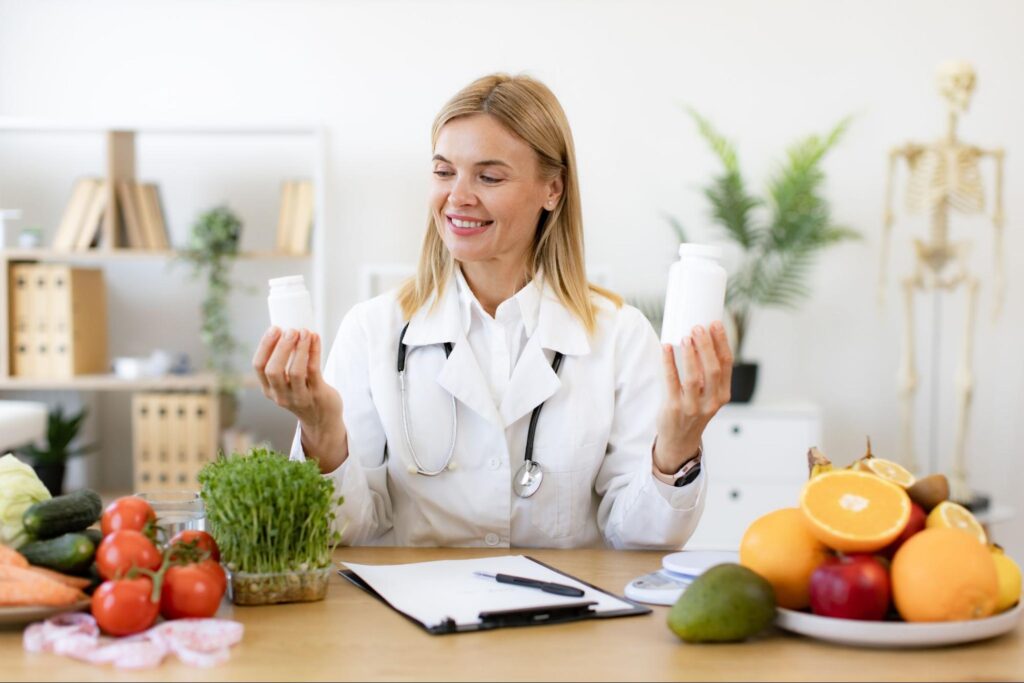 A smiling Caucasian functional medicine practitioner in a lab coat shows pill bottles made from natural ingredients, with fruits and vegetables on the table.