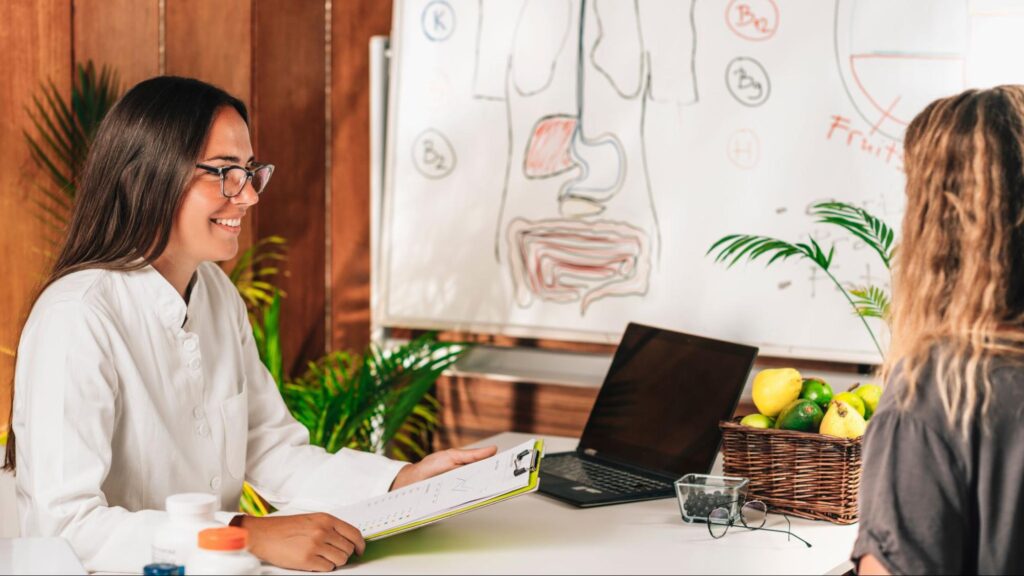 A female client consults a functional medicine practitioner to discuss health insights. A human anatomy chart and fresh fruits are visible in the background.