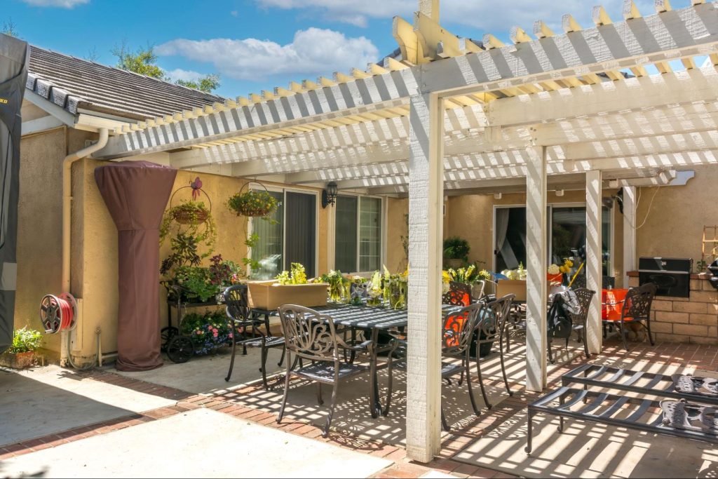 A backyard patio with tables, chairs, and a barbecue grill is surrounded by decorative plants. Glass windows and doors are visible in the background.