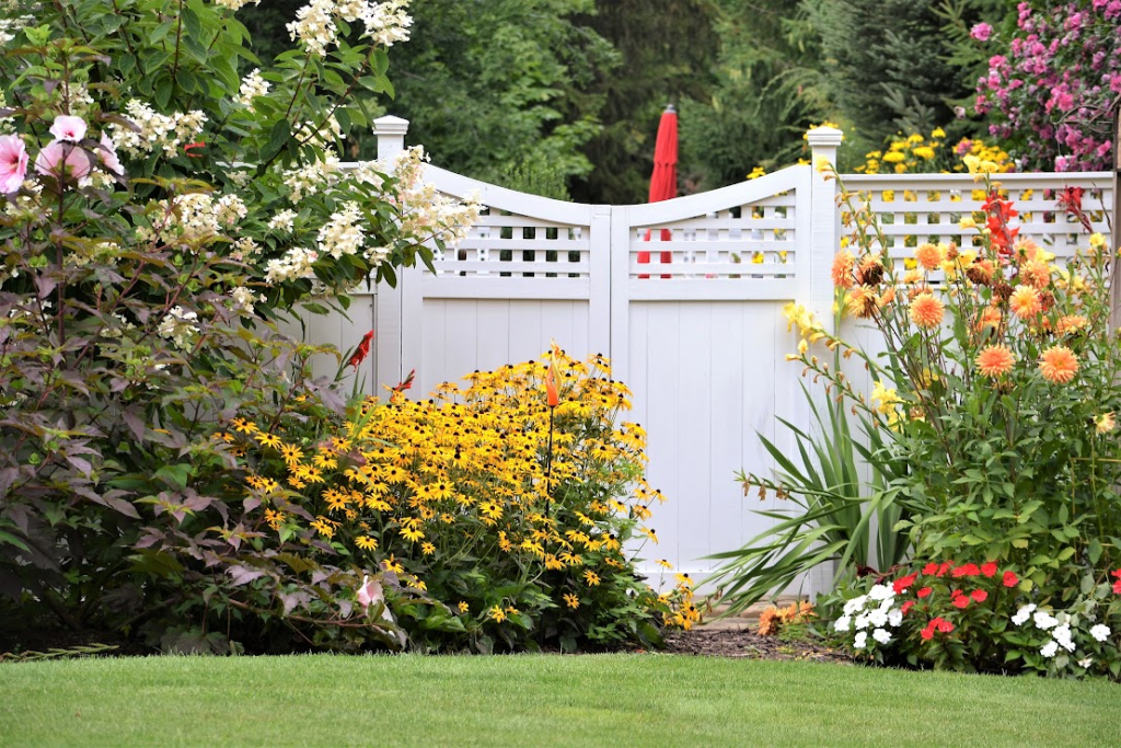 A colorful flower garden is against a white vinyl fence.