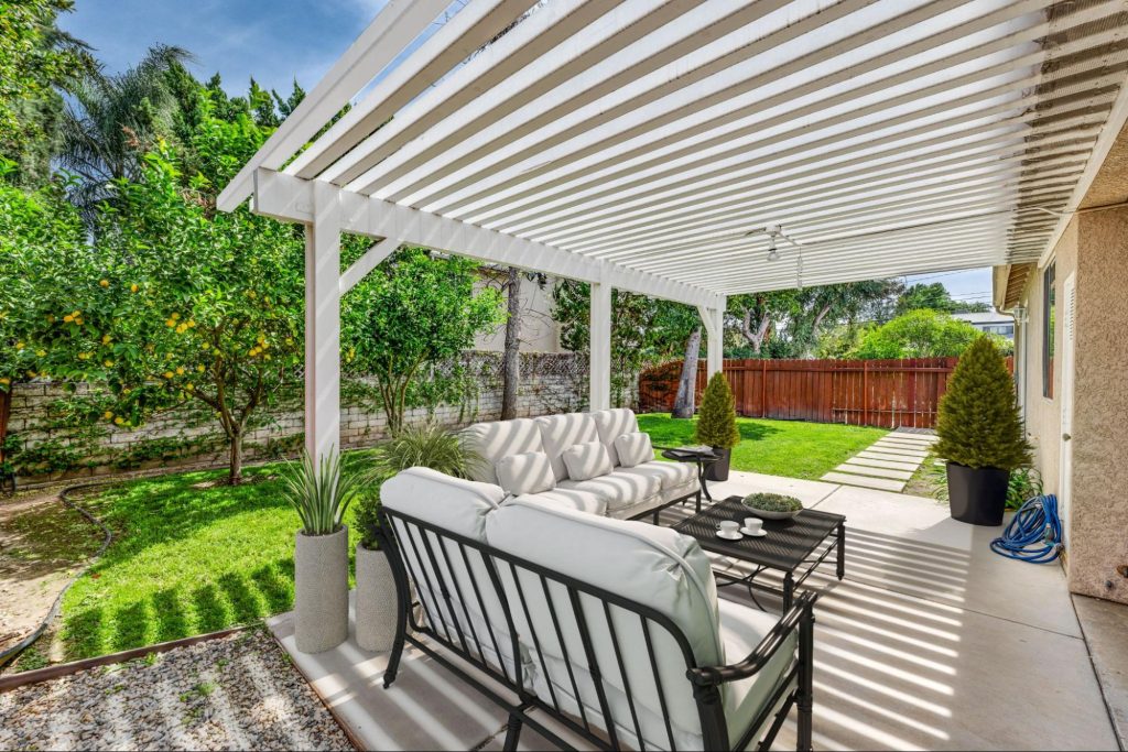 A sunny patio with a 3D-decorated house featuring a stylish vinyl patio cover with a slatted design for shade and elegance.