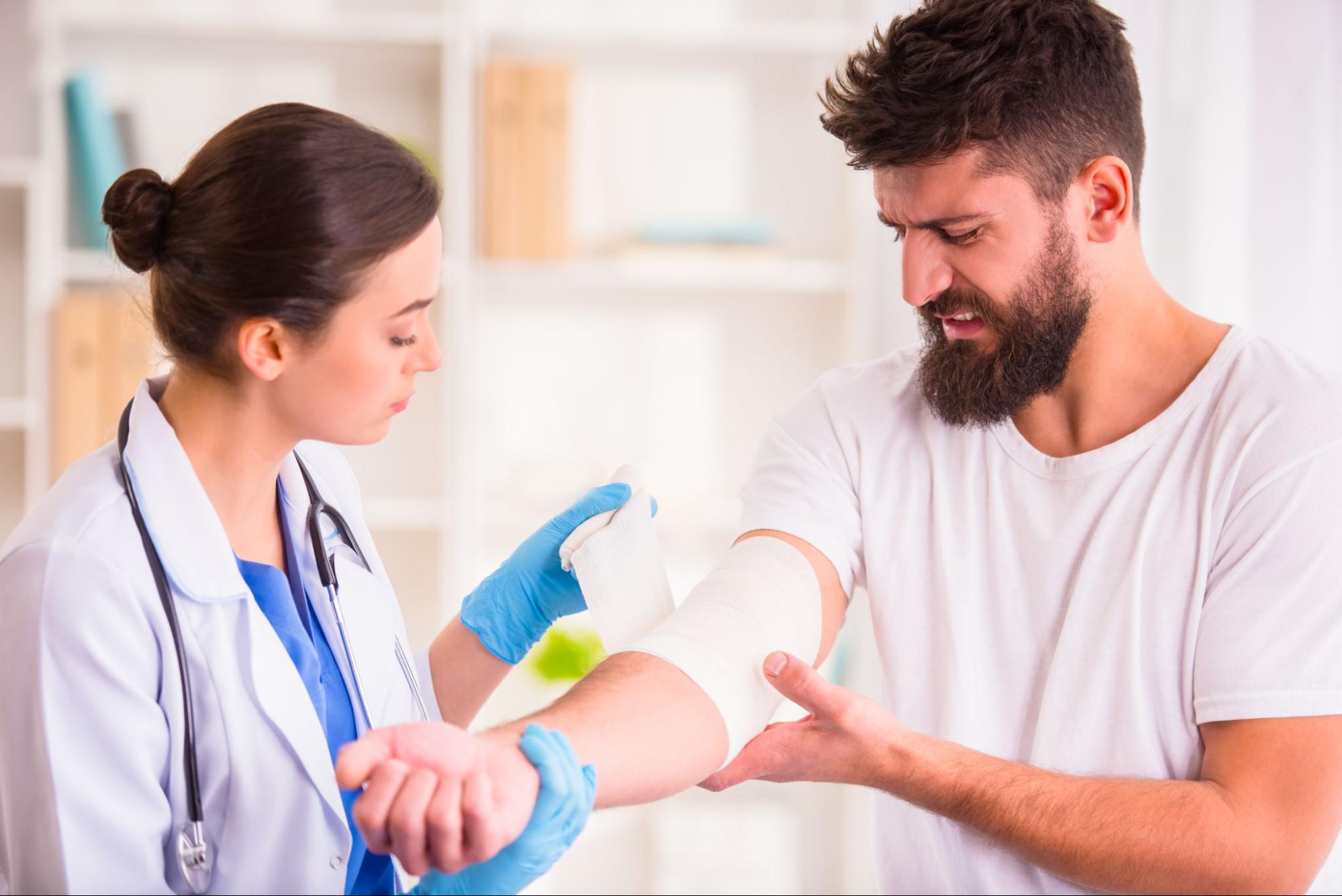 A female doctor wearing blue scrubs and a white jacket wrapping a bandage around a wound on a man’s arm.