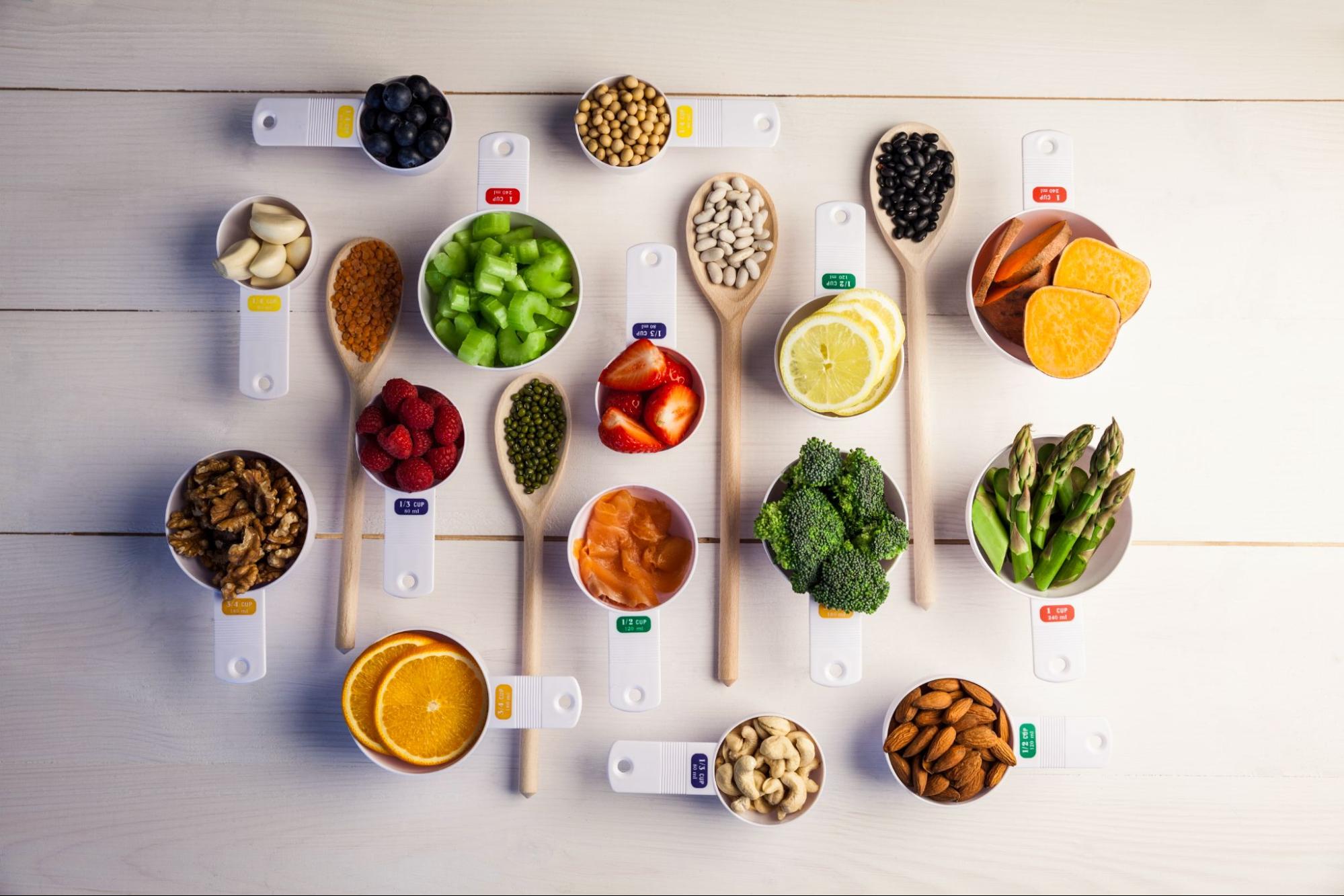Portion cups and spoons of healthy foods and ingredients, such as fruit, vegetables, and nuts, are on a wooden table.