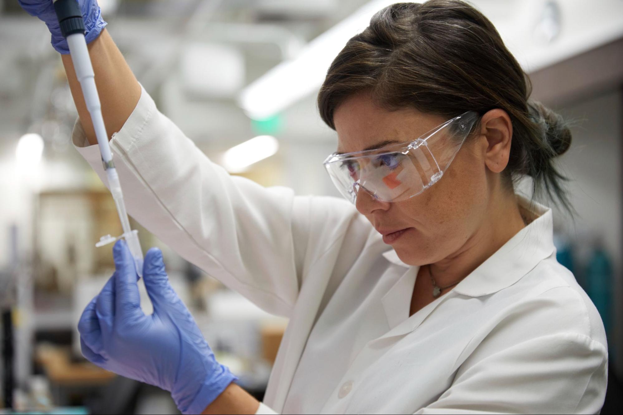 A female scientist wearing safety glasses and blue medical gloves using a pipette for a sample in a test tube.
