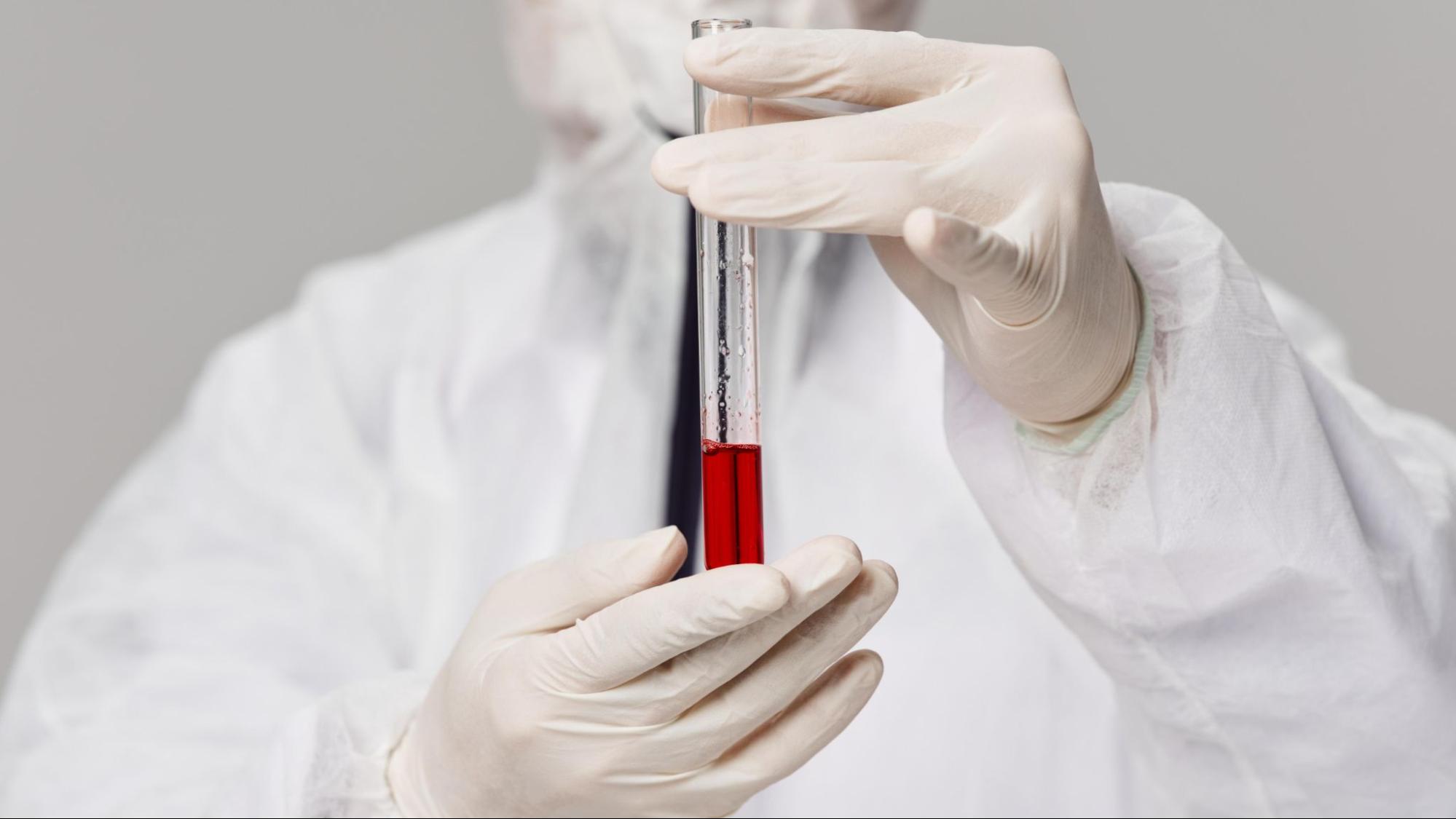 A scientist in full PPE and white gloves holding a vial of blood.