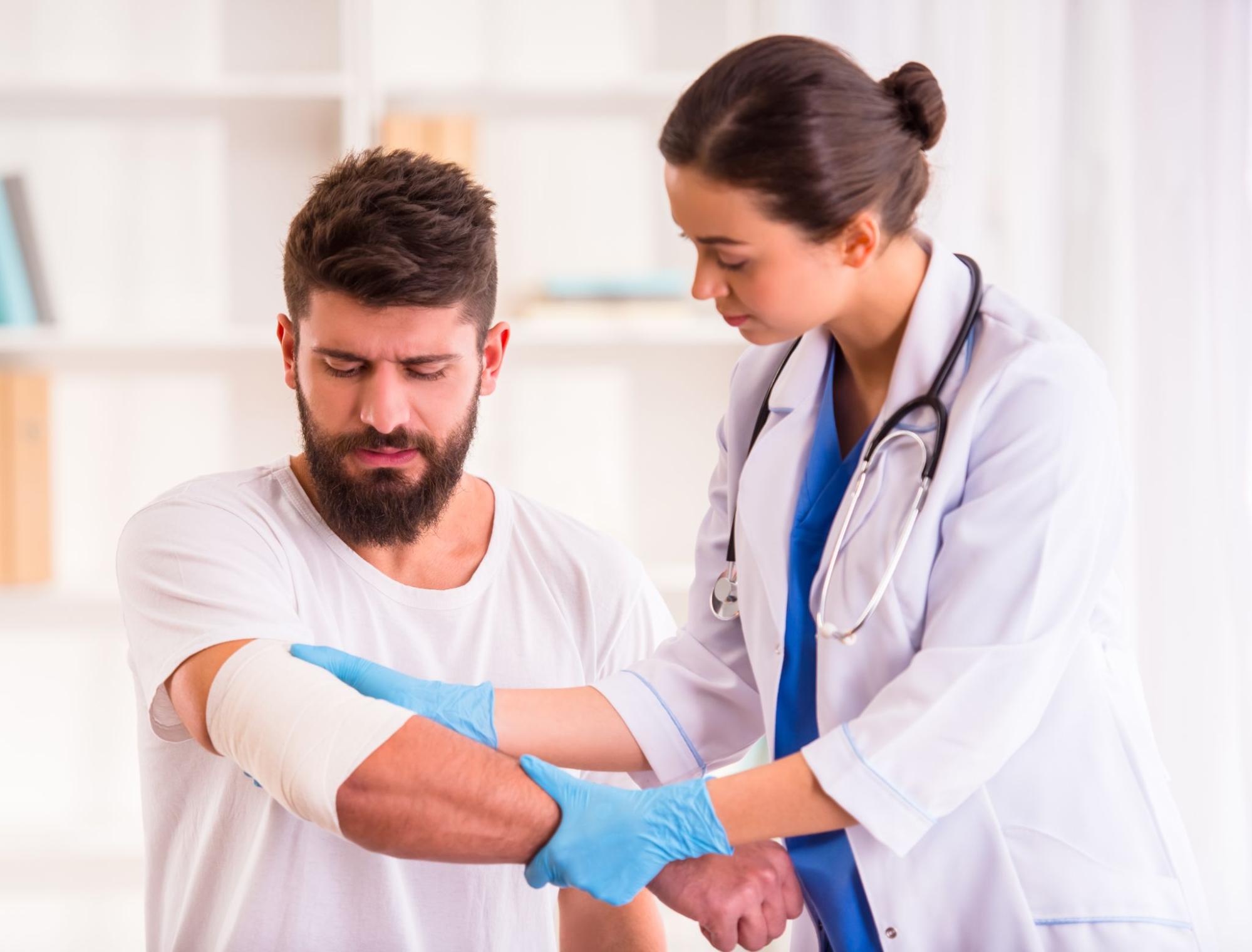 A female doctor wearing blue scrubs, a white coat, and a stethoscope bandaging a man’s elbow in a hospital to treat pressure ulcer symptoms.