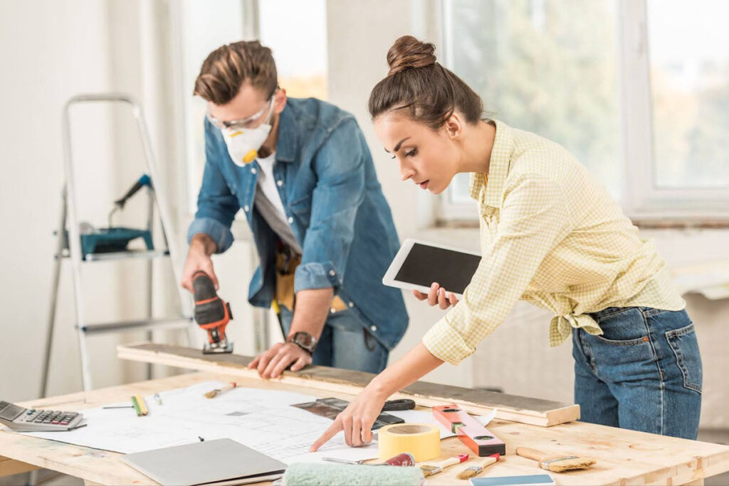 A man uses an electric sander on a wooden plank in the background while his partner checks their progress against home improvement ideas on her tablet.